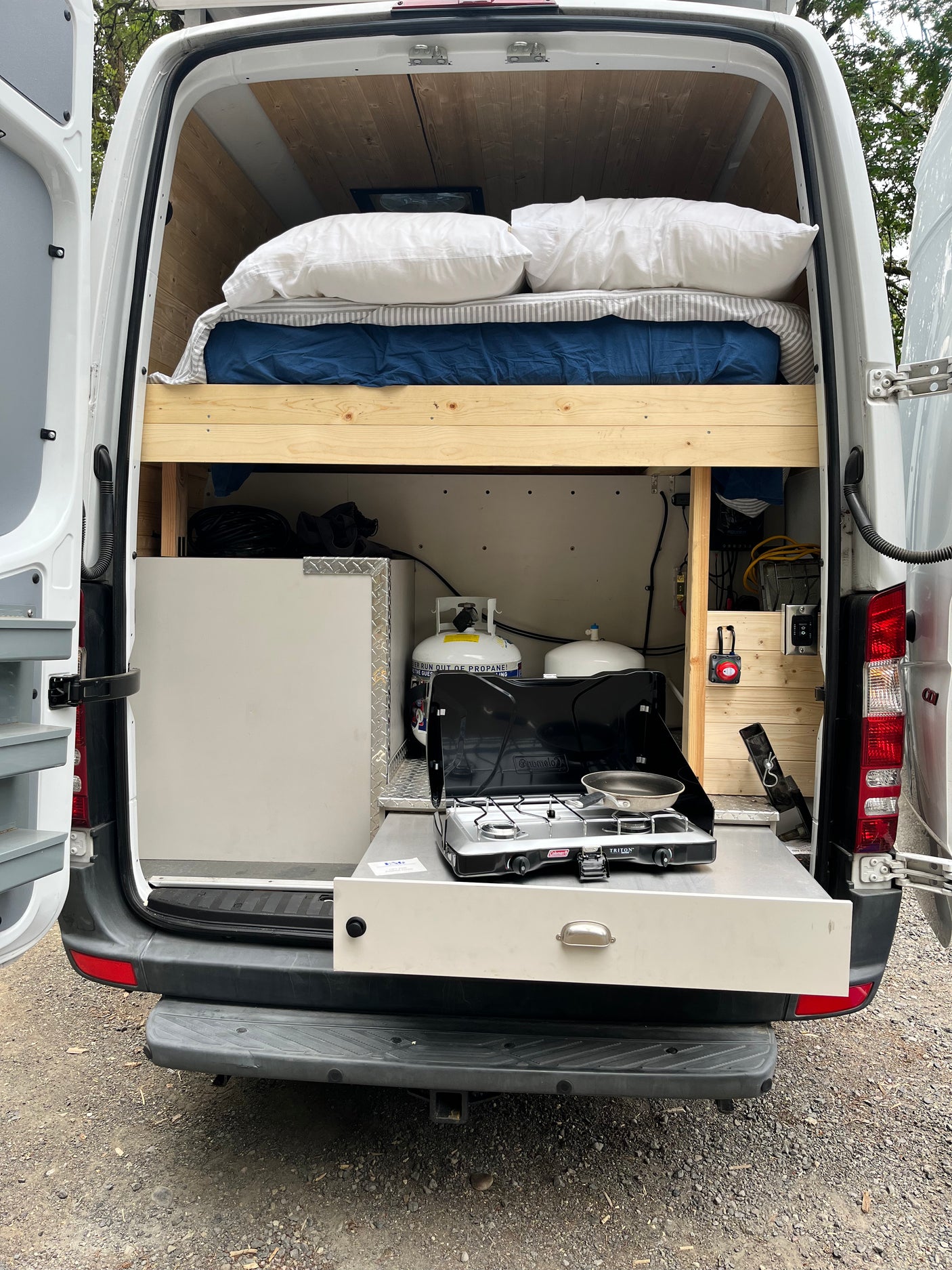Rear view of an RV rental camper van showing a comfy bed on the left and a compact kitchen with stove & propane.