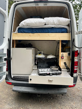 Rear view of an RV rental camper van showing a comfy bed on the left and a compact kitchen with stove & propane.