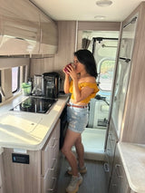 Woman sips a drink in her modern RV rental kitchen, featuring a compact stovetop and light wood cabinets. Perfect for road trips!