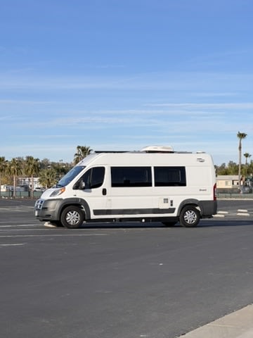 A white class B RV rental, resembling a large camper van, is parked alone in a sunny, empty asphalt lot with palm trees in the distance.