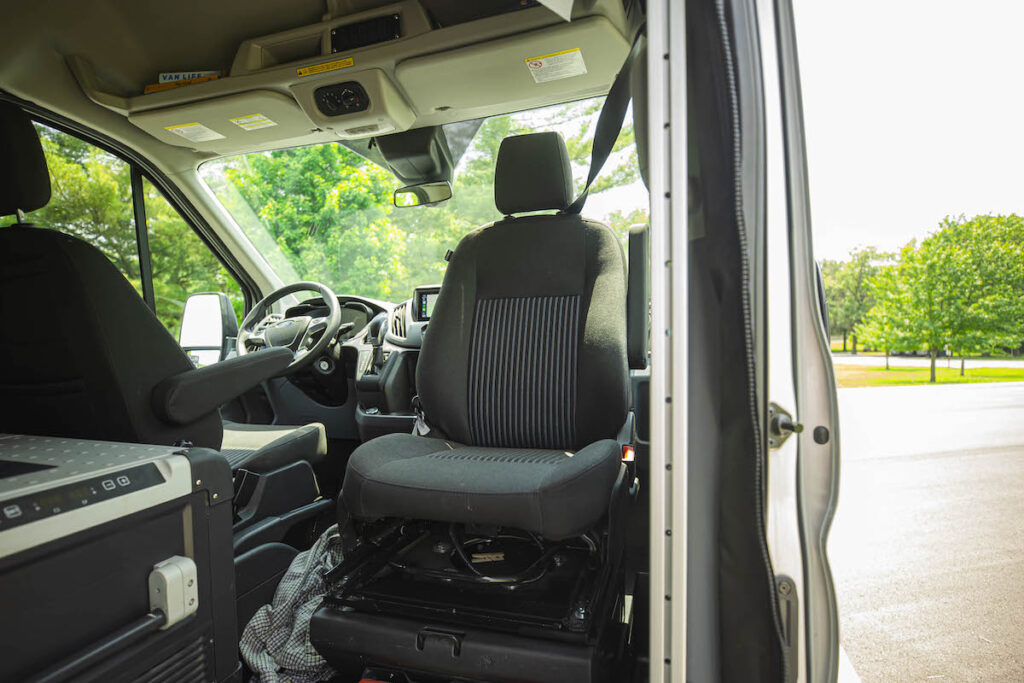 Interior of an rv rental van showing the swivel passenger seat and front cabin. Trees visible through the windshield.