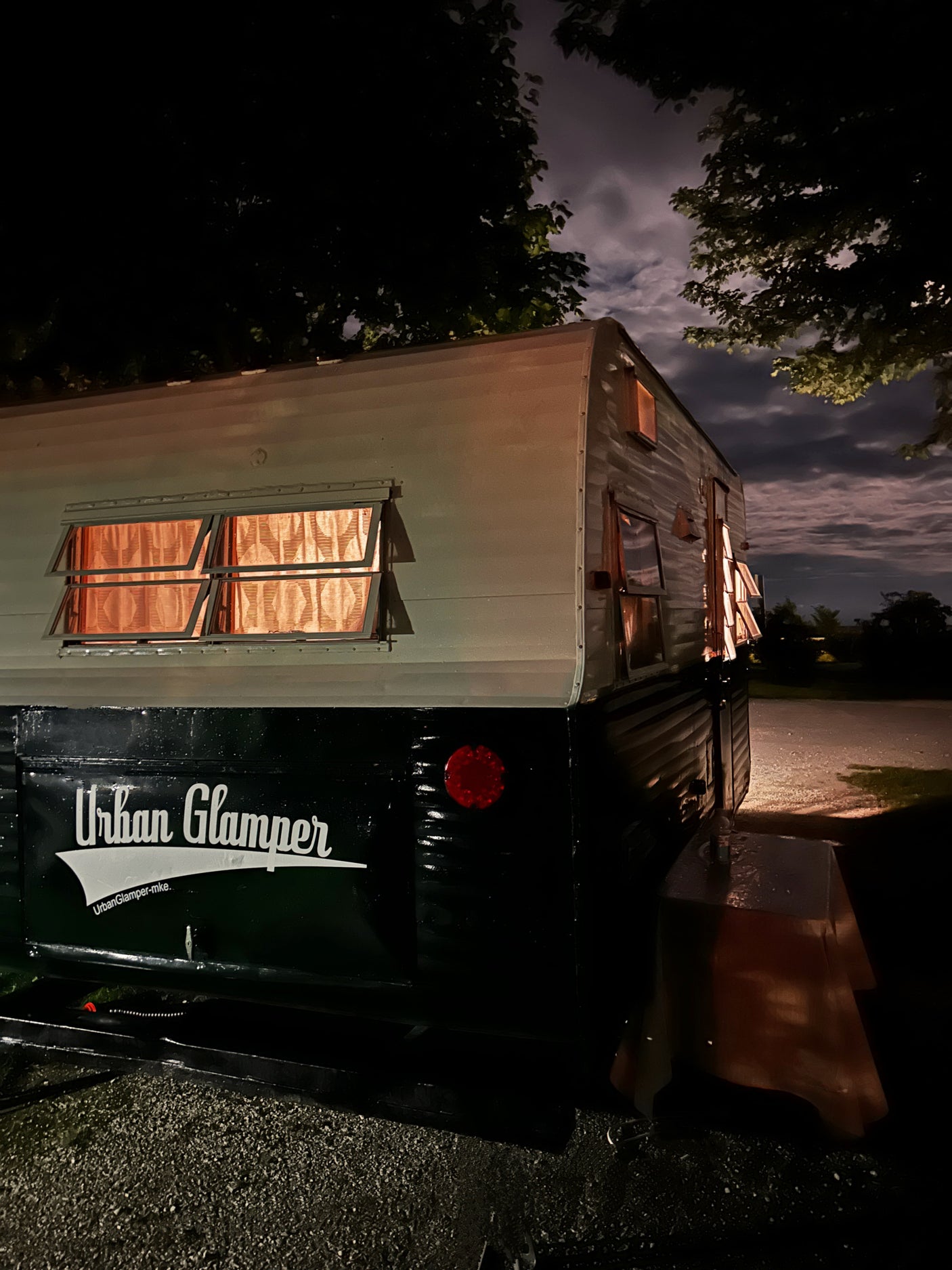 Cozy vintage 'Urban Glamper' RV rental at night, glowing warmly from within. Perfect for glamping under the cloudy sky.
