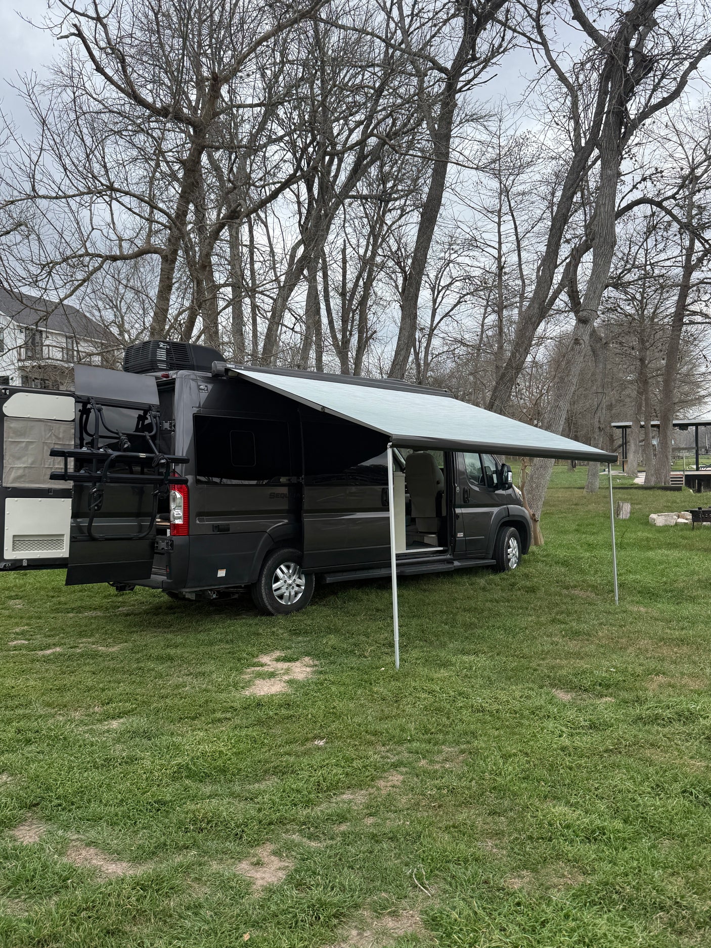 Dark camper van RV rental with an extended awning, parked on green grass among bare trees on a cloudy day.