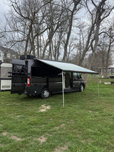 Dark camper van RV rental with an extended awning, parked on green grass among bare trees on a cloudy day.