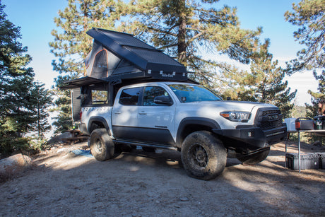 White Toyota Tacoma with an open Alu-Cab truck camper tent set up at a scenic campsite. An ideal RV rental alternative.