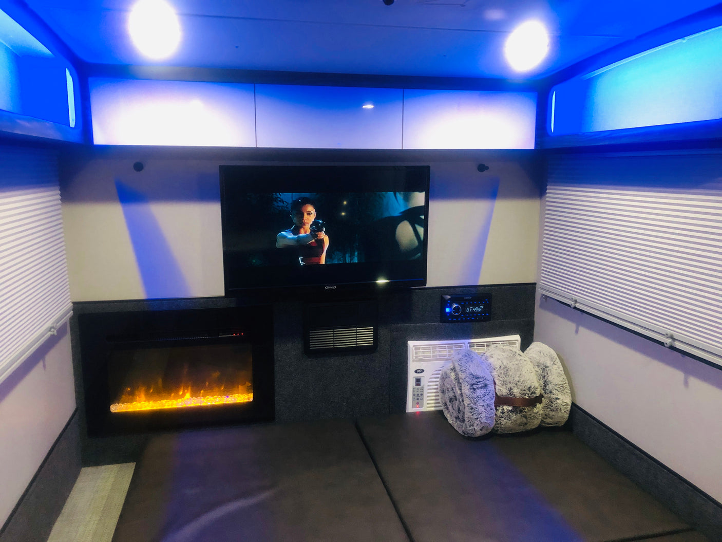 An RV rental's cozy interior, featuring a TV, electric fireplace, AC unit, and soft bedding, all illuminated by cool blue lights.