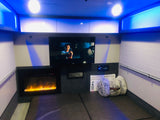 An RV rental's cozy interior, featuring a TV, electric fireplace, AC unit, and soft bedding, all illuminated by cool blue lights.