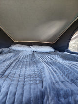 Cozy pop-top RV rental bed. A comfortable quilted grey blanket and two white pillows are ready for rest. Trees outside a mesh window.
