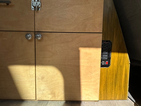 RV rental interior with custom wooden cabinets, power inverter (AC/USB outlets). Sunlight streams in.