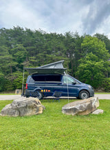 A blue camper van with pop-top & awning, like an RV rental, is set up with chairs & table on grass by a forest.