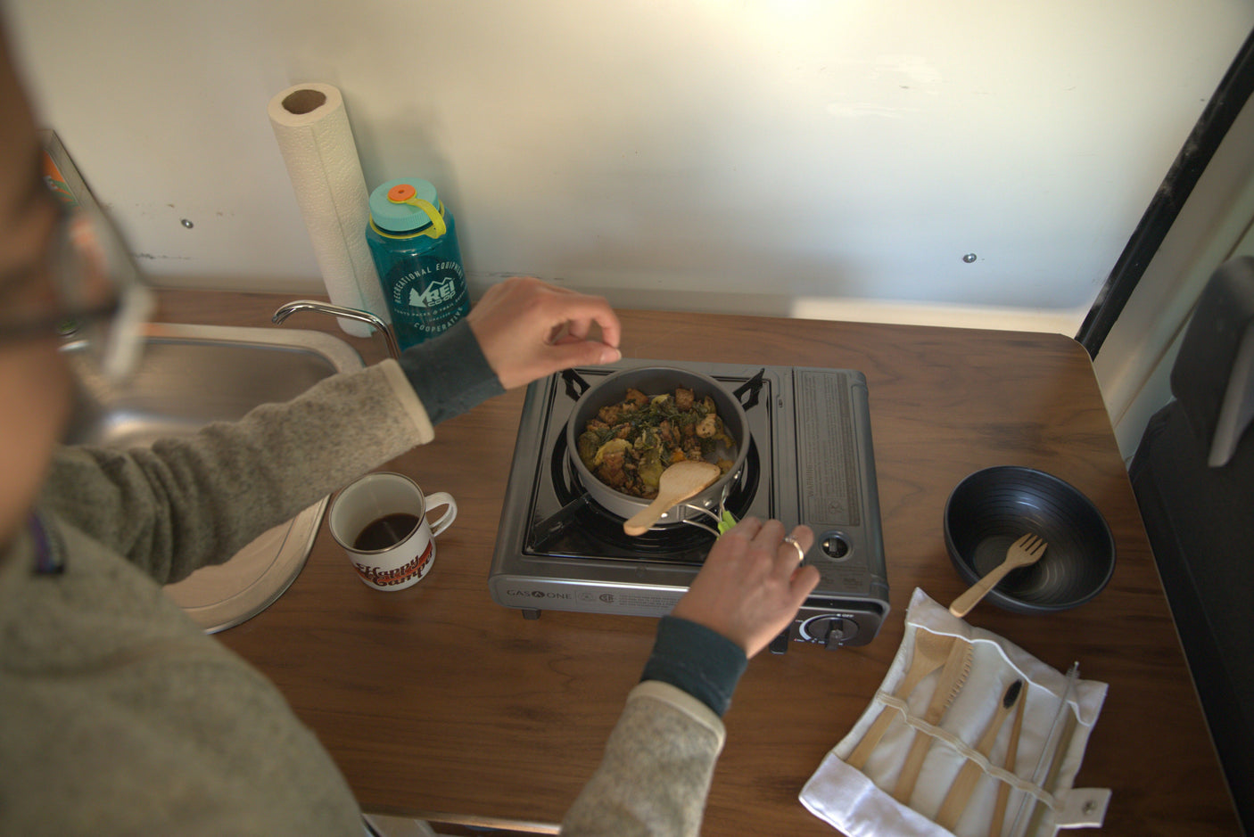 Preparing food on a gas stove in a compact rv rental kitchen, with a sink, water bottle, and 'Happy Camper' mug visible.