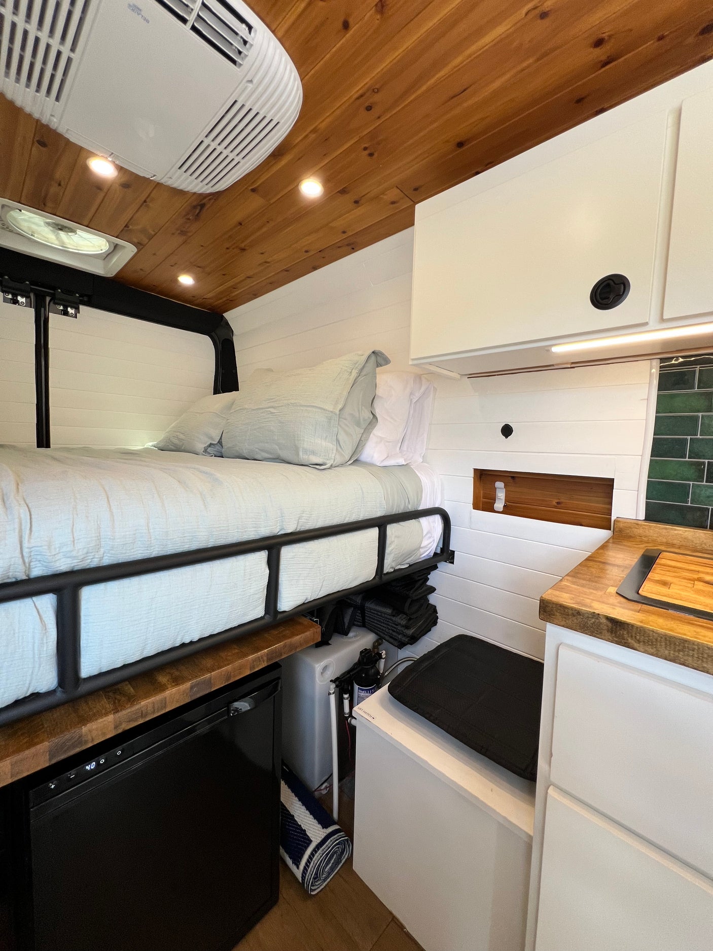 RV rental interior: cozy bed, small kitchen with wood counters, refrigerator, water system, and overhead storage.