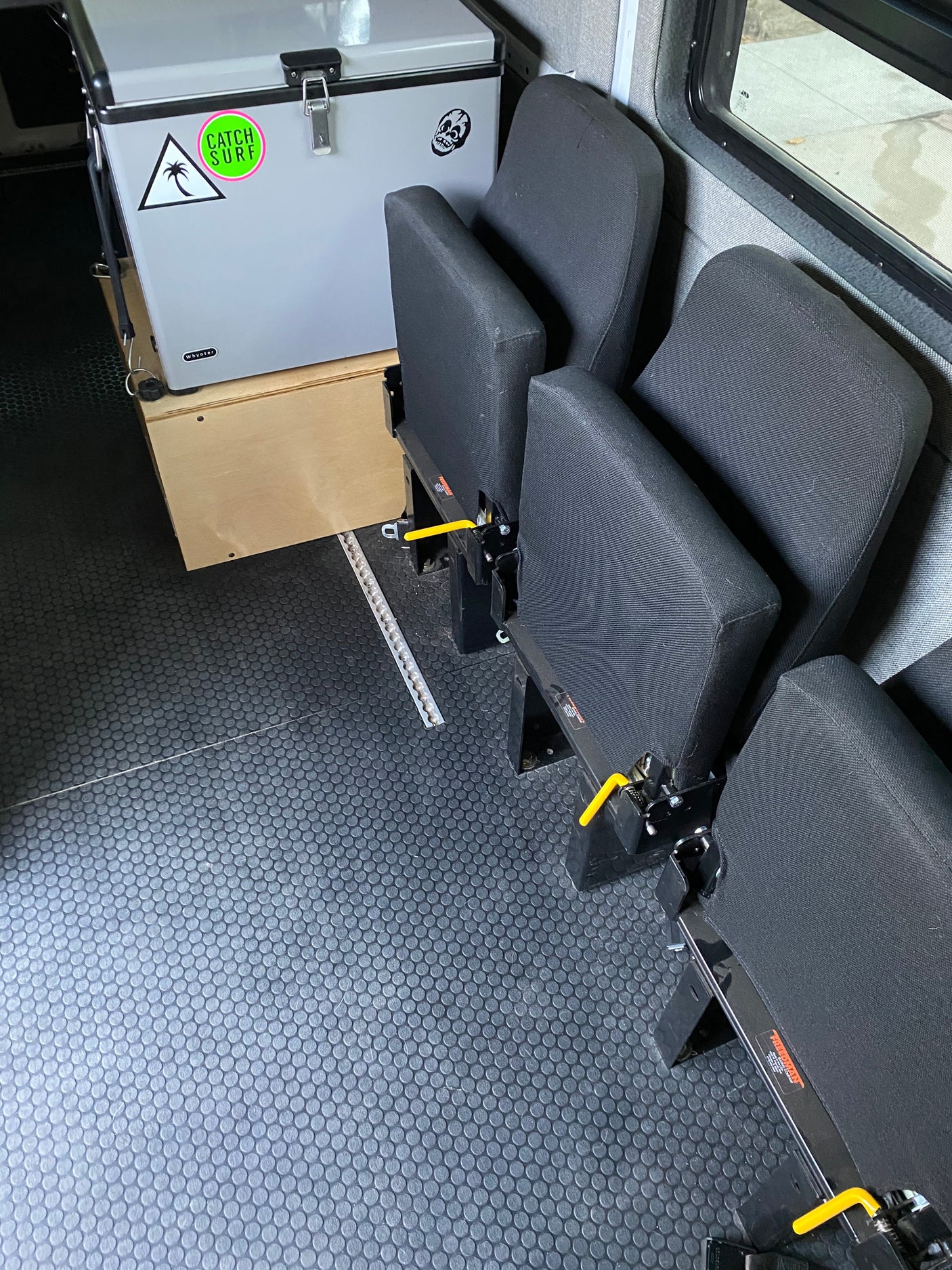 RV rental interior: black modular seats on floor tracks, durable textured floor, and a cooler on a wooden base.