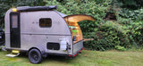 Teardrop RV rental parked in lush greenery, showing its open rear galley kitchen and side features.