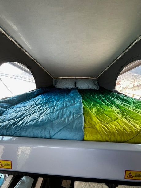 Inside an RV rental, a comfortable pop-top bed with vibrant blue and green sleeping bags, pillows, and bright windows.