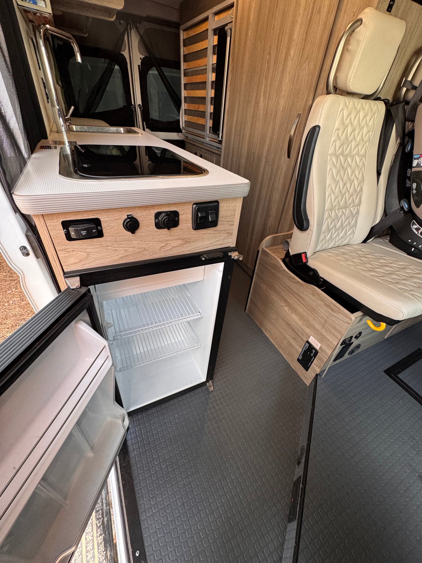 Inside this RV rental: a cozy kitchenette with a sink, cooktop, and an open mini-fridge. Features modern seating and wood-grain interior.