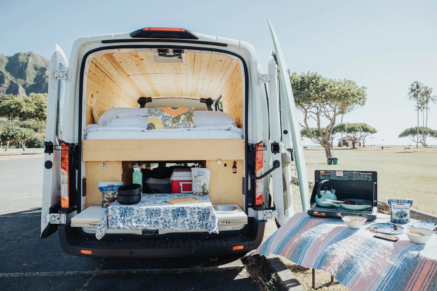 An RV rental's rear opens to a cozy bed & pull-out kitchen. Outdoor cooking on a picnic table completes this camper van setup.