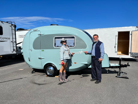 A smiling couple stands with their light blue, retro-style Barefoot travel trailer, perfect for an RV rental adventure.