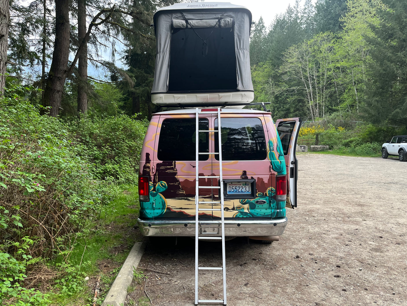 Rear view of a colorful rv rental van with a rooftop tent open & ladder down, parked in a lush forest in WA.