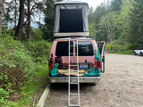 Rear view of a colorful rv rental van with a rooftop tent open & ladder down, parked in a lush forest in WA.
