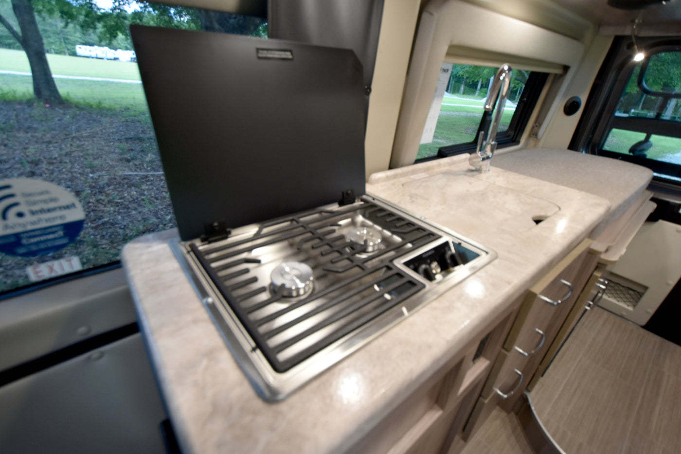An RV rental interior, showing a kitchen counter with a two-burner stove, sink, and views of a green outdoor area through windows. — photo 12