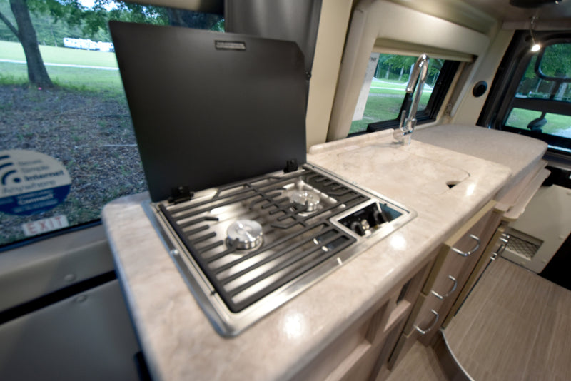 An RV rental interior, showing a kitchen counter with a two-burner stove, sink, and views of a green outdoor area through windows. — photo 12