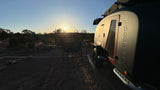 RV rental adventure: a sunlit teardrop camper and double chair at a desert campsite as the sun rises over the horizon.