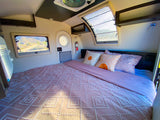 Inside an RV rental: a cozy bedroom with pink geometric bedding, scenic windows, and overhead storage for travel comfort.