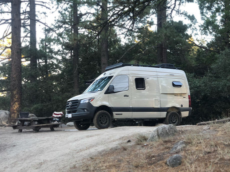 Beige Winnebago Revel Sprinter RV rental parked at a rustic campsite with a picnic table, ready for adventure.