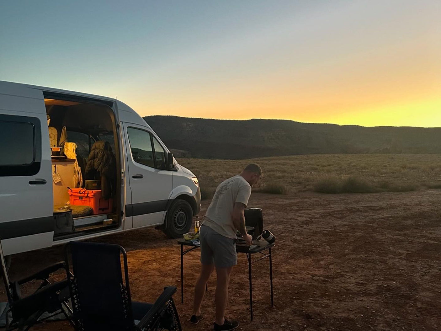 Golden sunset: A man cooks outside his white RV rental van, glowing interior visible, enjoying the vast outdoor landscape.