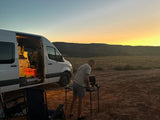 Golden sunset: A man cooks outside his white RV rental van, glowing interior visible, enjoying the vast outdoor landscape.