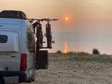 Revel 4x4 RV rental with bikes parked lakeside at a hazy sunset. Golden sun reflects on calm water, hills in distance.
