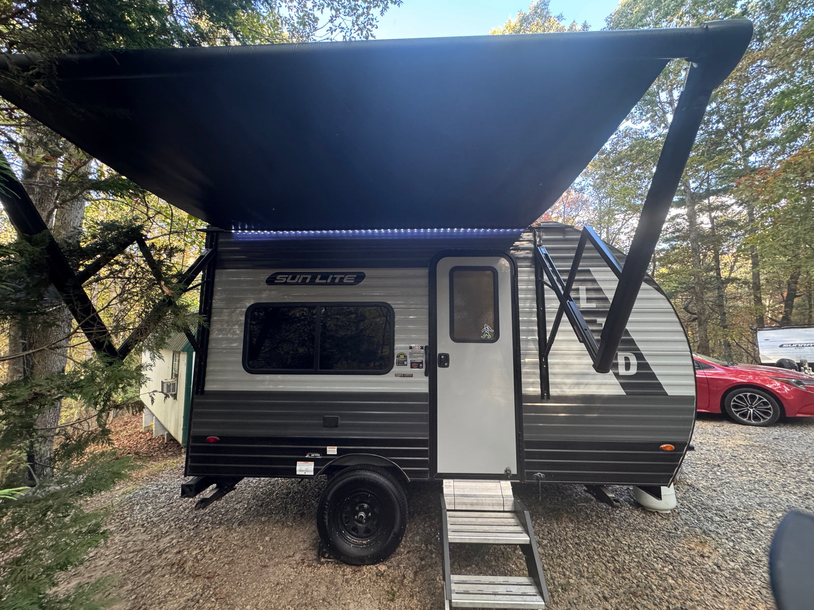 Enjoy this RV rental! A Sun Lite travel trailer with an illuminated awning is set up on gravel in a wooded camp spot.