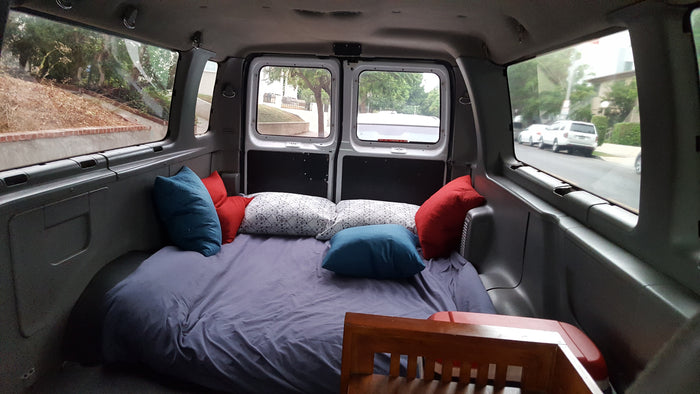 Cozy RV rental interior with a full bed & pillows. Views out side & back windows of parked cars & trees.