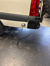 RV rental camper van's rear: red taillight, spare tire, & external power cable connected. Parked on a dark, worn floor.