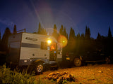 RV rental camping scene: A travel trailer, pickup truck, and campfire ring under a starry night sky in the forest.