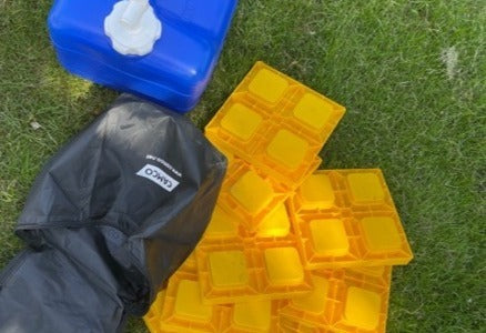 RV rental essentials: Blue water jug, yellow leveling blocks, and black Camco gear bag on grass for camping.