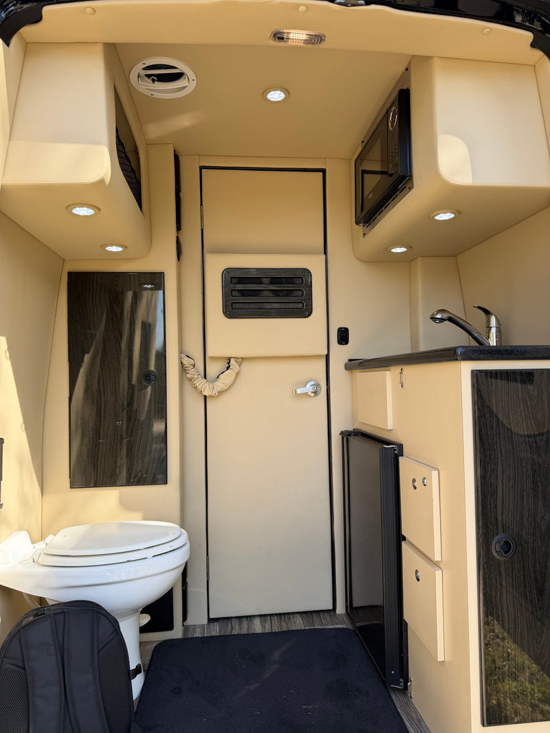 Modern RV rental interior: beige cabinetry, dark wood accents. Features compact kitchen with sink, microwave, fridge, and toilet. — photo 3