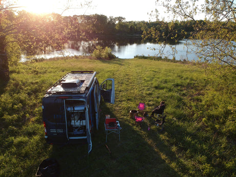 An RV rental camper van parked by a serene lake at sunset, with a person relaxing in a chair, ideal for camping.