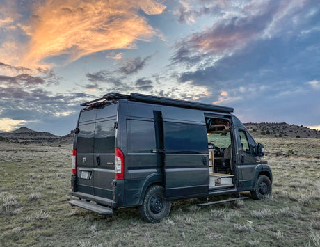 Explore in this black RV rental camper van, parked in a scenic field under a vibrant sunset sky. Adventure awaits!