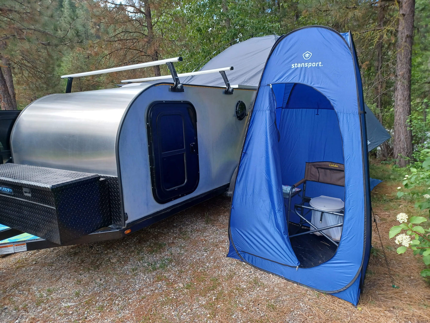 An RV rental teardrop trailer with roof racks, next to a blue Stansport privacy tent housing a portable toilet for camping.