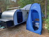 An RV rental teardrop trailer with roof racks, next to a blue Stansport privacy tent housing a portable toilet for camping.