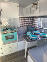 Cozy vintage RV rental interior featuring a turquoise stove, dining set with blue bowls, and plaid curtains.
