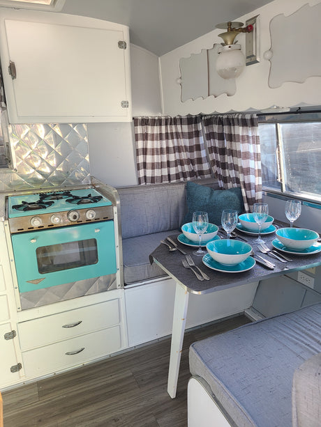 Cozy vintage RV rental interior featuring a turquoise stove, dining set with blue bowls, and plaid curtains.