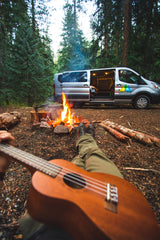 First-person view of camping: legs, ukulele, and a bright campfire. An RV rental van awaits in the lush forest.