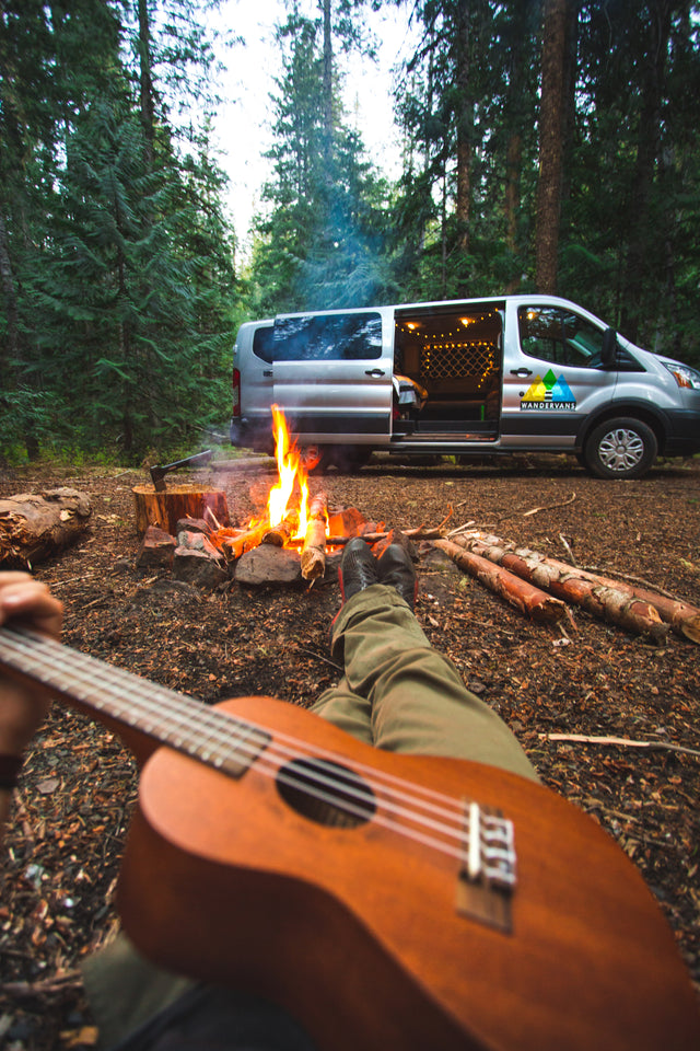 First-person view of camping: legs, ukulele, and a bright campfire. An RV rental van awaits in the lush forest.
