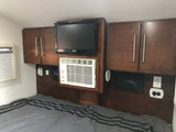 Cozy RV rental bedroom featuring a wall-mounted TV, compact AC unit, cabinets, and a charging center above a striped bed.