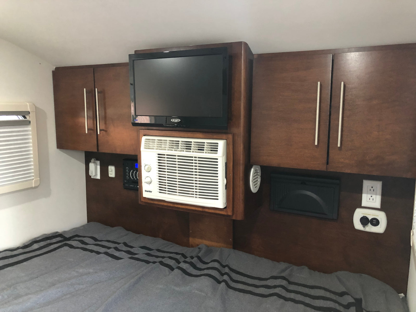Cozy RV rental bedroom featuring a wall-mounted TV, compact AC unit, cabinets, and a charging center above a striped bed.