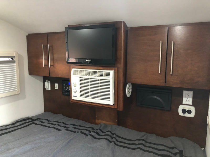 Cozy RV rental bedroom featuring a wall-mounted TV, compact AC unit, cabinets, and a charging center above a striped bed. — photo 4