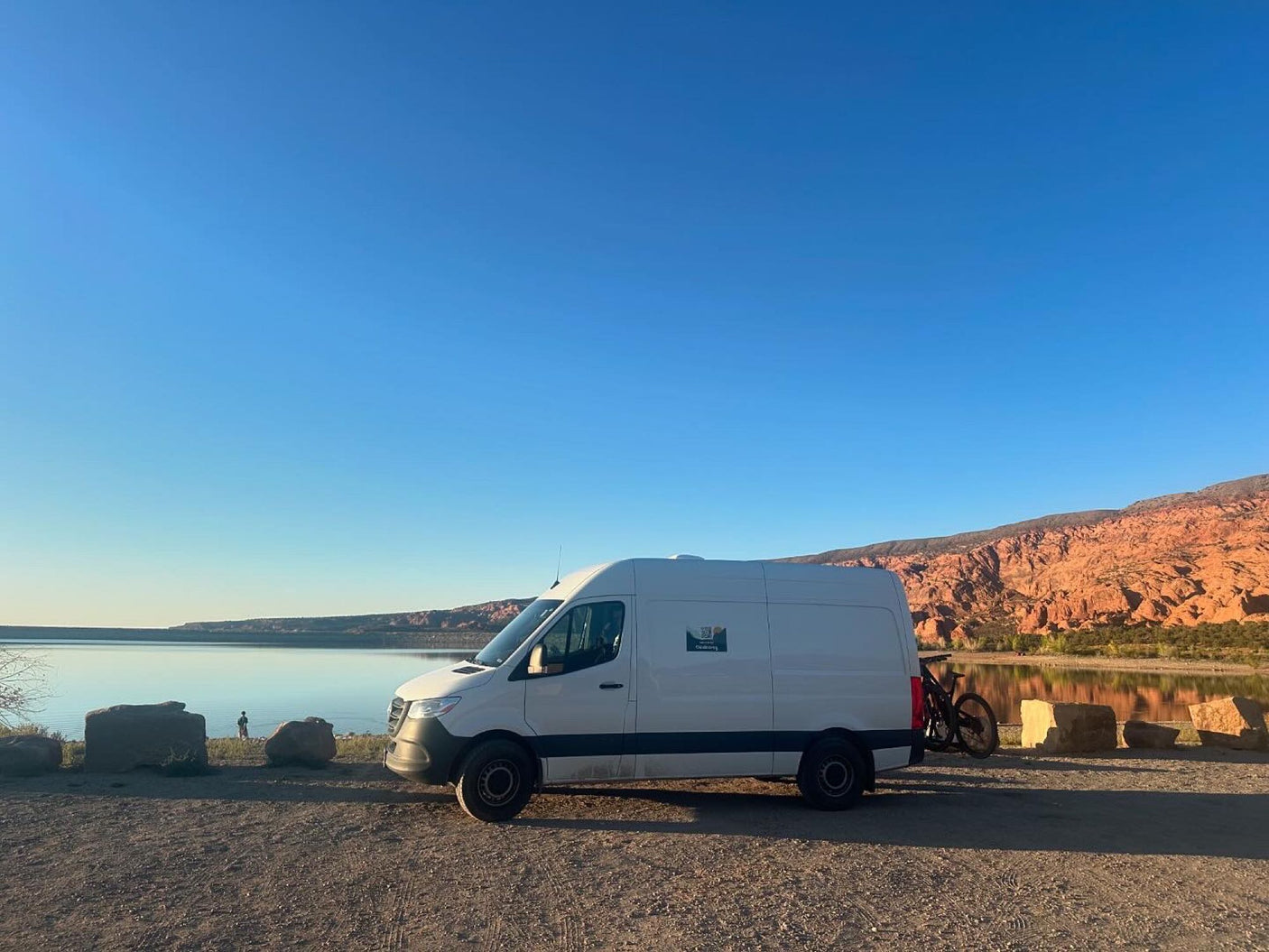 Discover adventure with this RV rental camper van parked by a serene lake & red rock cliffs, ready for biking.